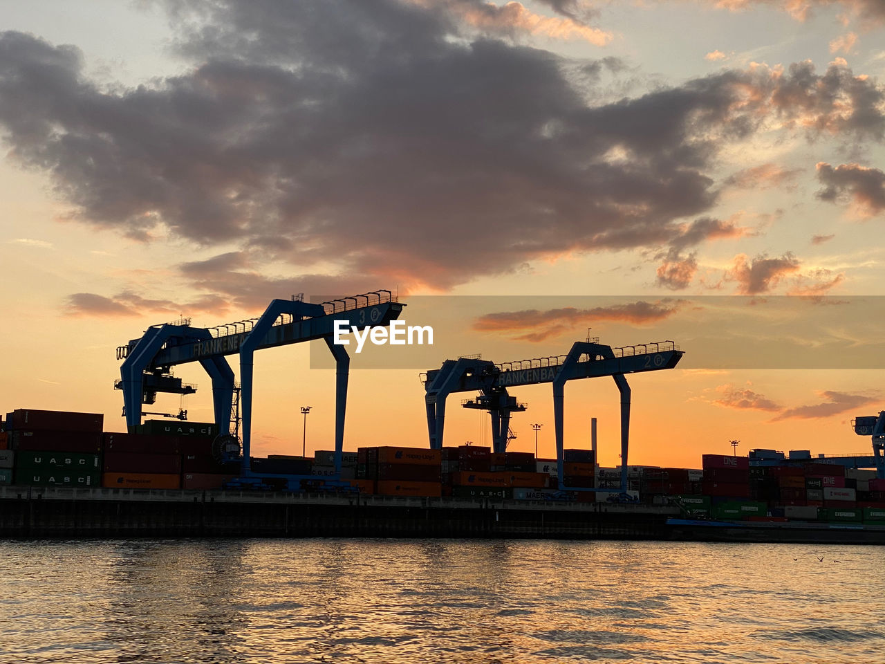 SILHOUETTE CRANES AT RIVERBANK AGAINST SKY AT SUNSET