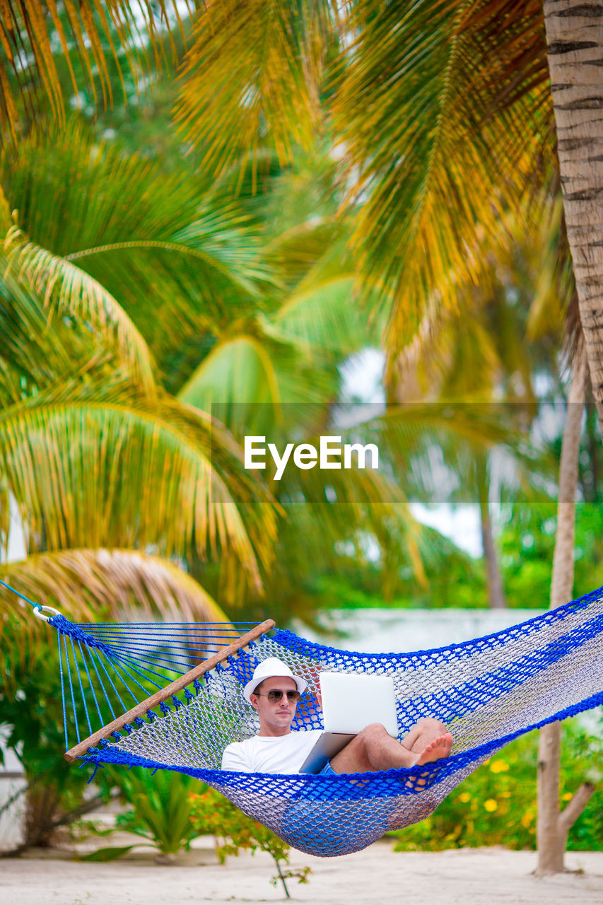 Man using laptop while relaxing in hammock against palm trees at beach