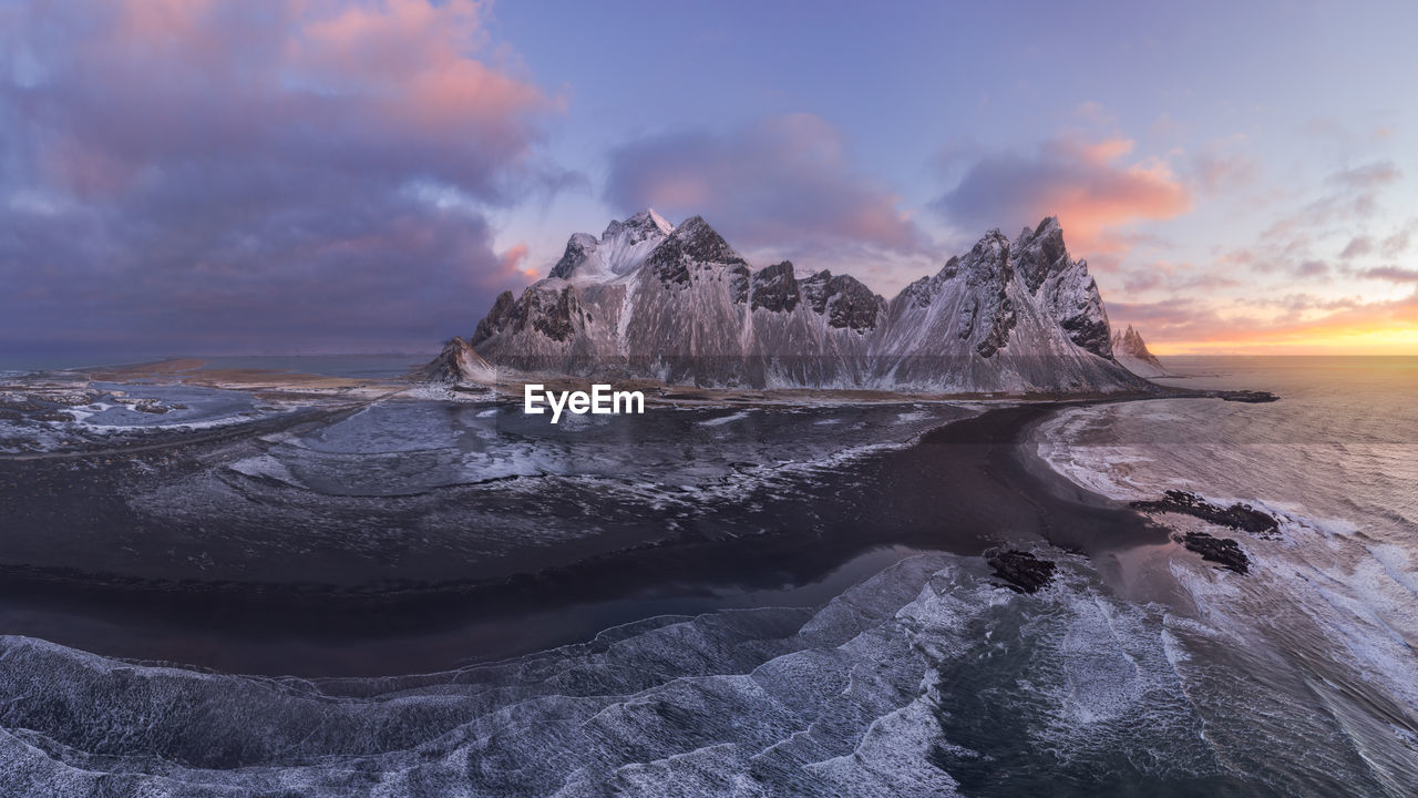 Drone view of calm frozen endless sea surrounding high rocky snowy vestrahorn mountain during picturesque sunset in stockness beach, iceland