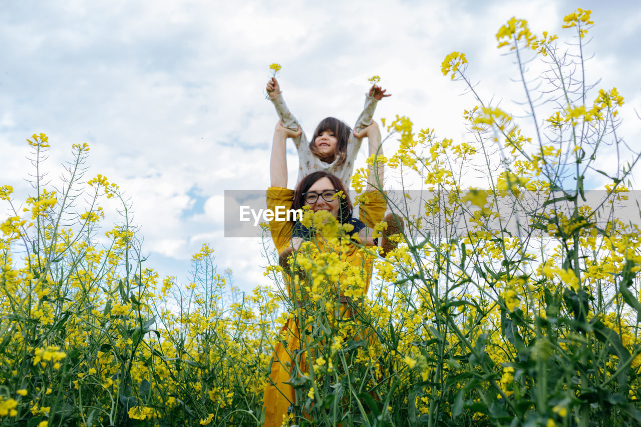 Smiling child sitting on mother shoulders in the field with yellow flowers against blue sky