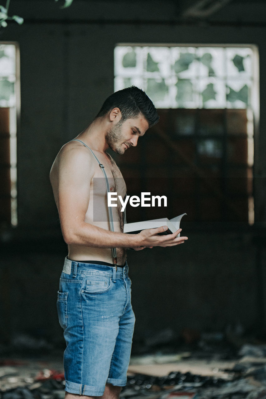 Side view of shirtless young man reading book standing at home