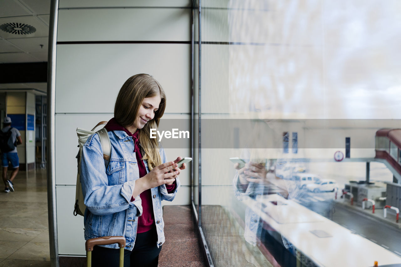 Young woman with suitcase using phone by glass window at airport