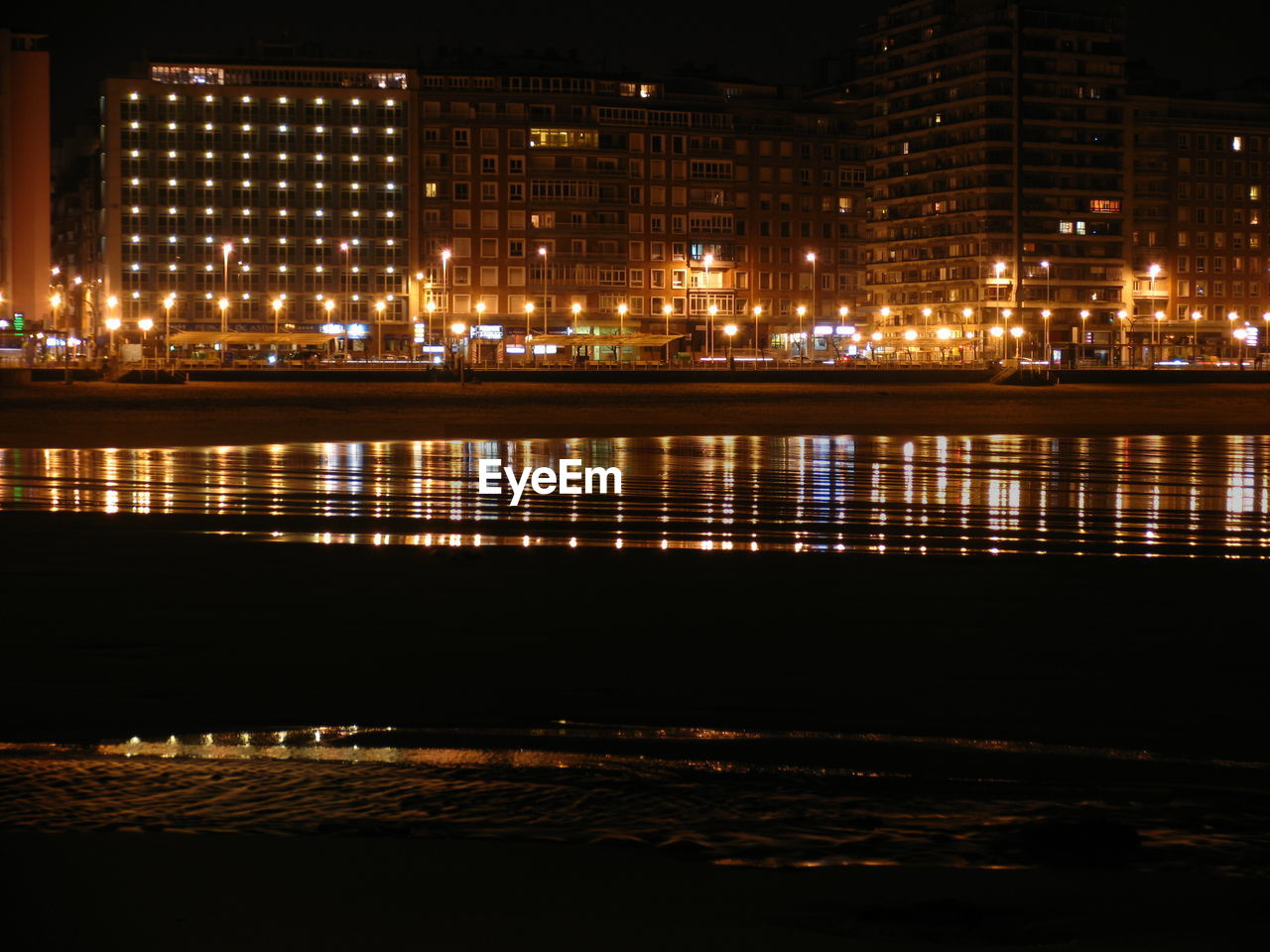 Illuminated buildings by beach against sky at night