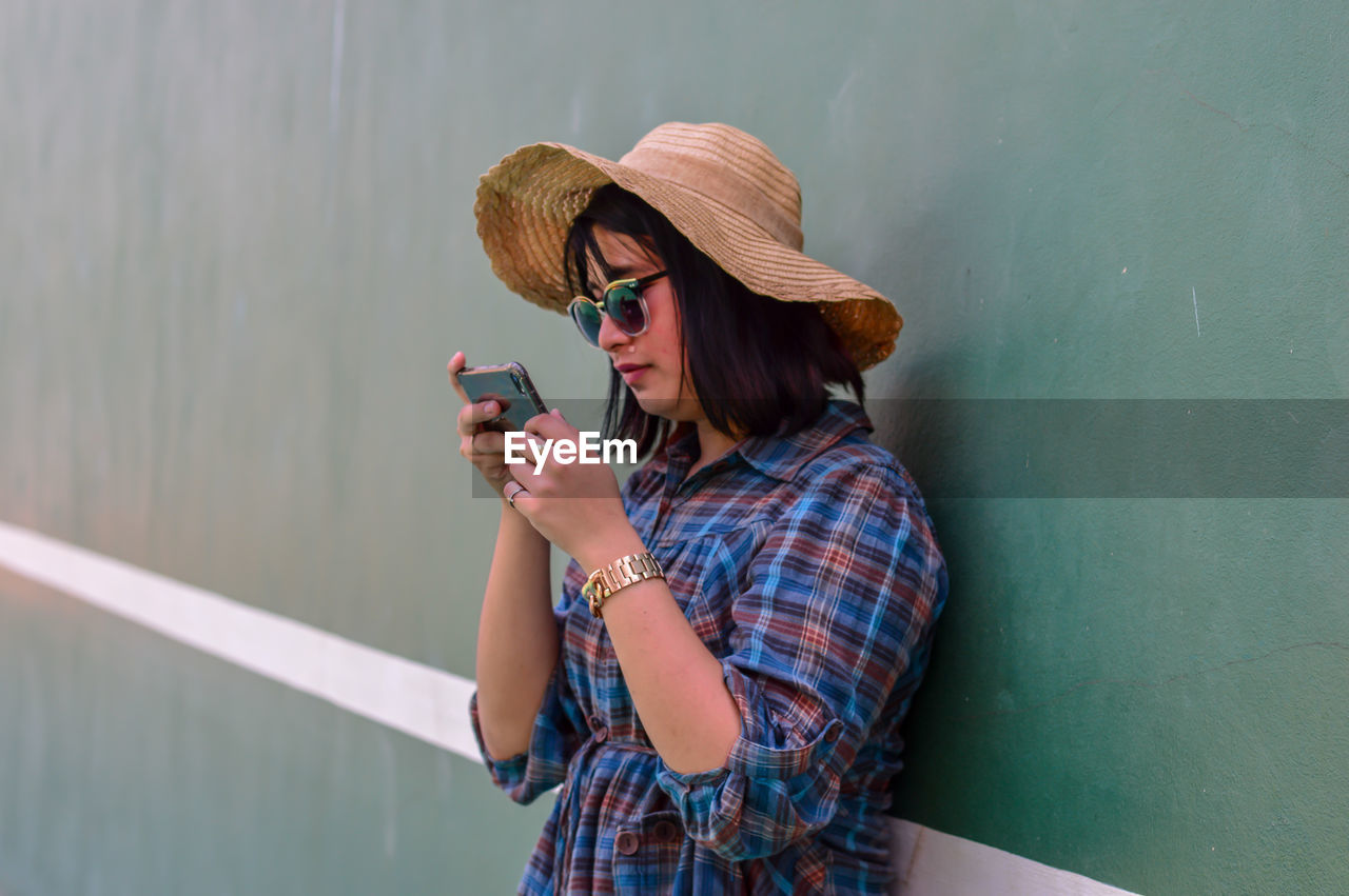 Woman wearing sunglasses and hat while using mobile phone against wall