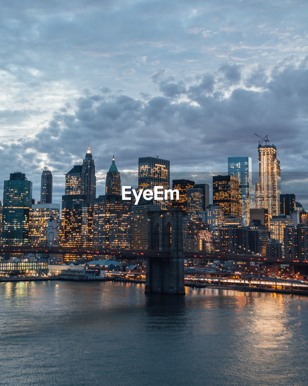 Brooklyn bridge over river by illuminated city against cloudy sky