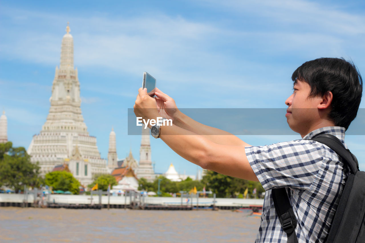 Side view of man photographing with mobile phone against sky