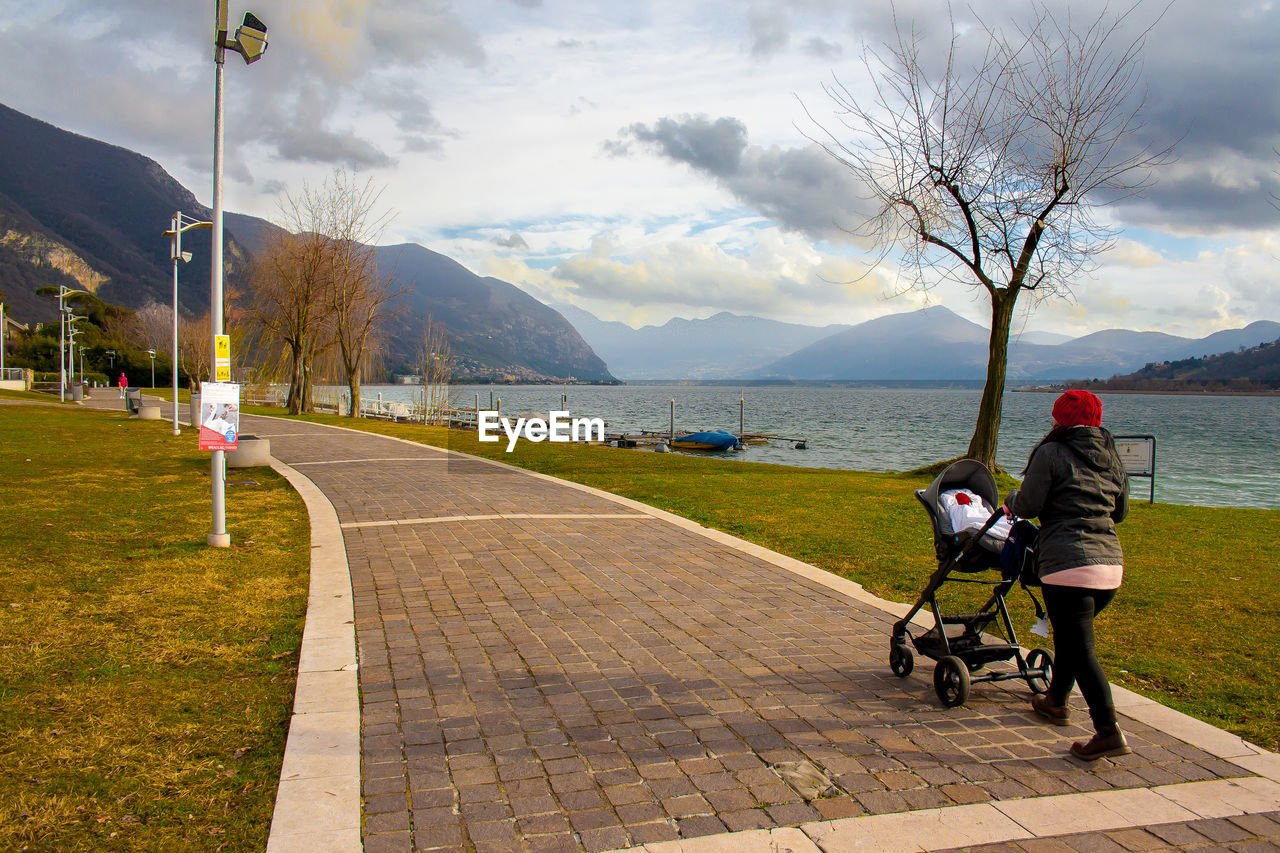 Rear view full length of woman pushing baby carriage by lake on footpath