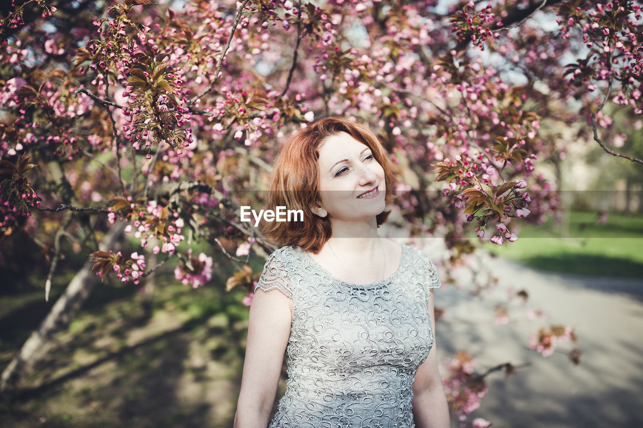 Happy middle aged armenian woman in an elegant dress under the blooming sakura tree. smiling