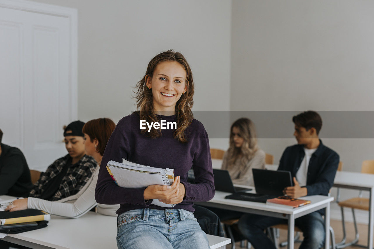 Portrait of happy young female student with book leaning on desk in classroom