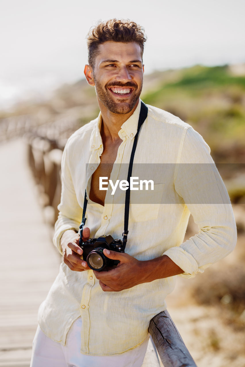 Smiling man holding camera against sky