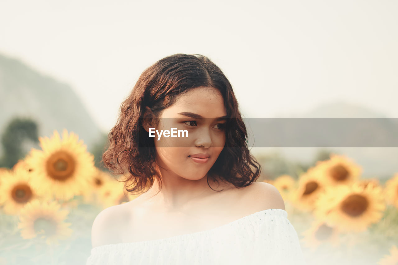 Close-up of thoughtful young woman standing at sunflower farm against clear sky