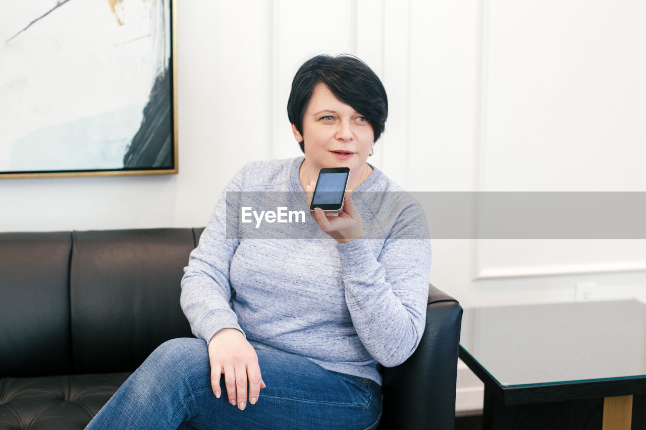 Middle age brunette woman sitting on couch sofa at home and talking on audio chat phone with friends