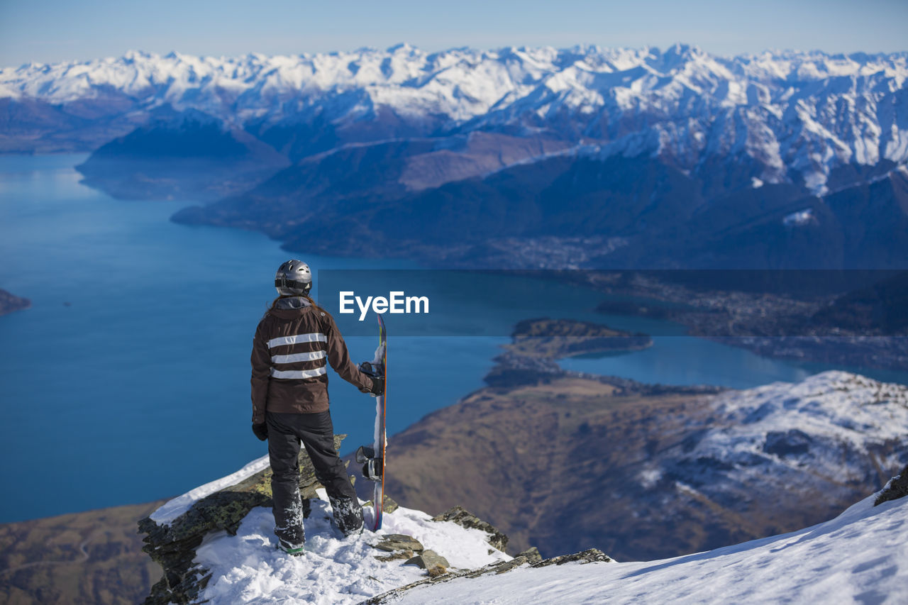 Rear view of person with snowboard standing on cliff by sea against mountains during winter