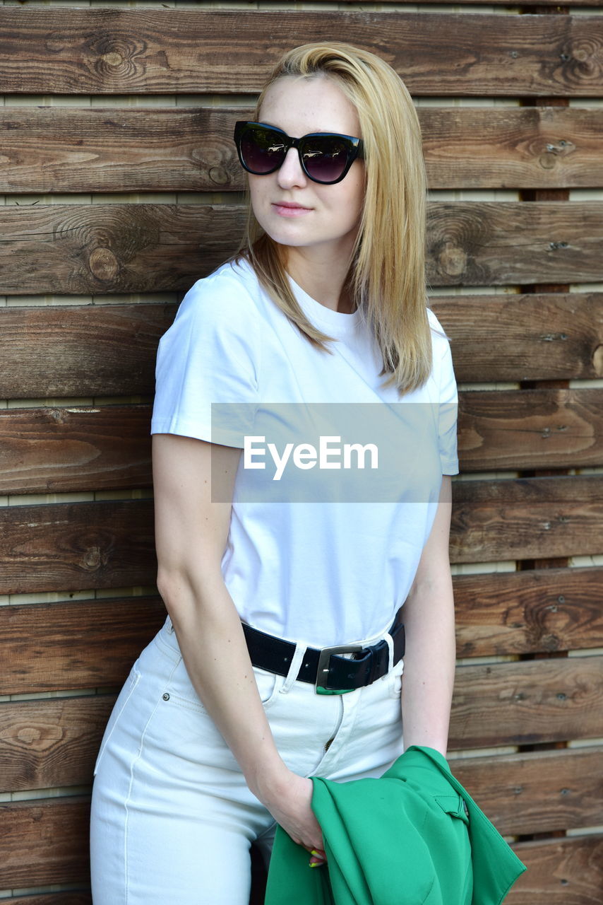 Portrait of young woman wearing sunglasses while standing against wall