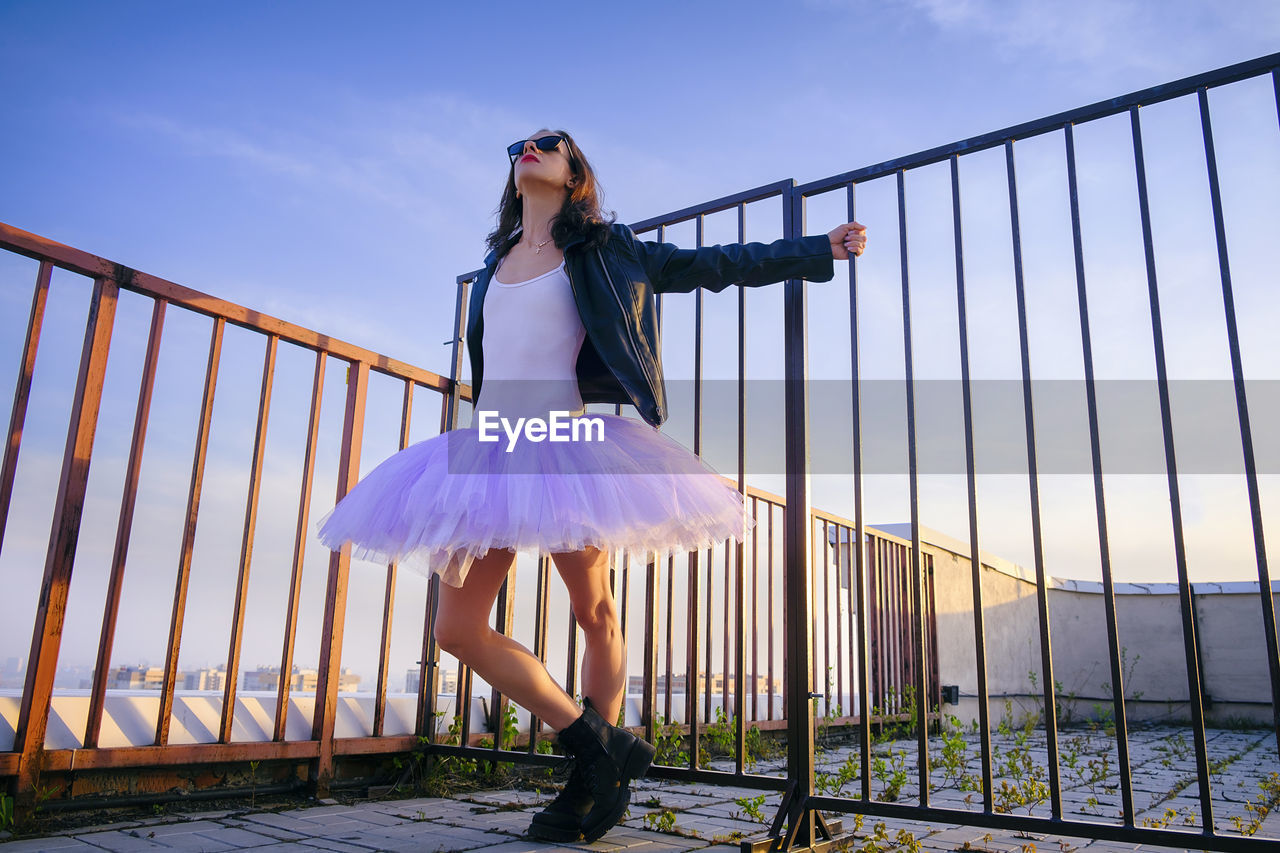 A ballerina in tutu stands at the fence on the roof at sunset in boots and a jacket