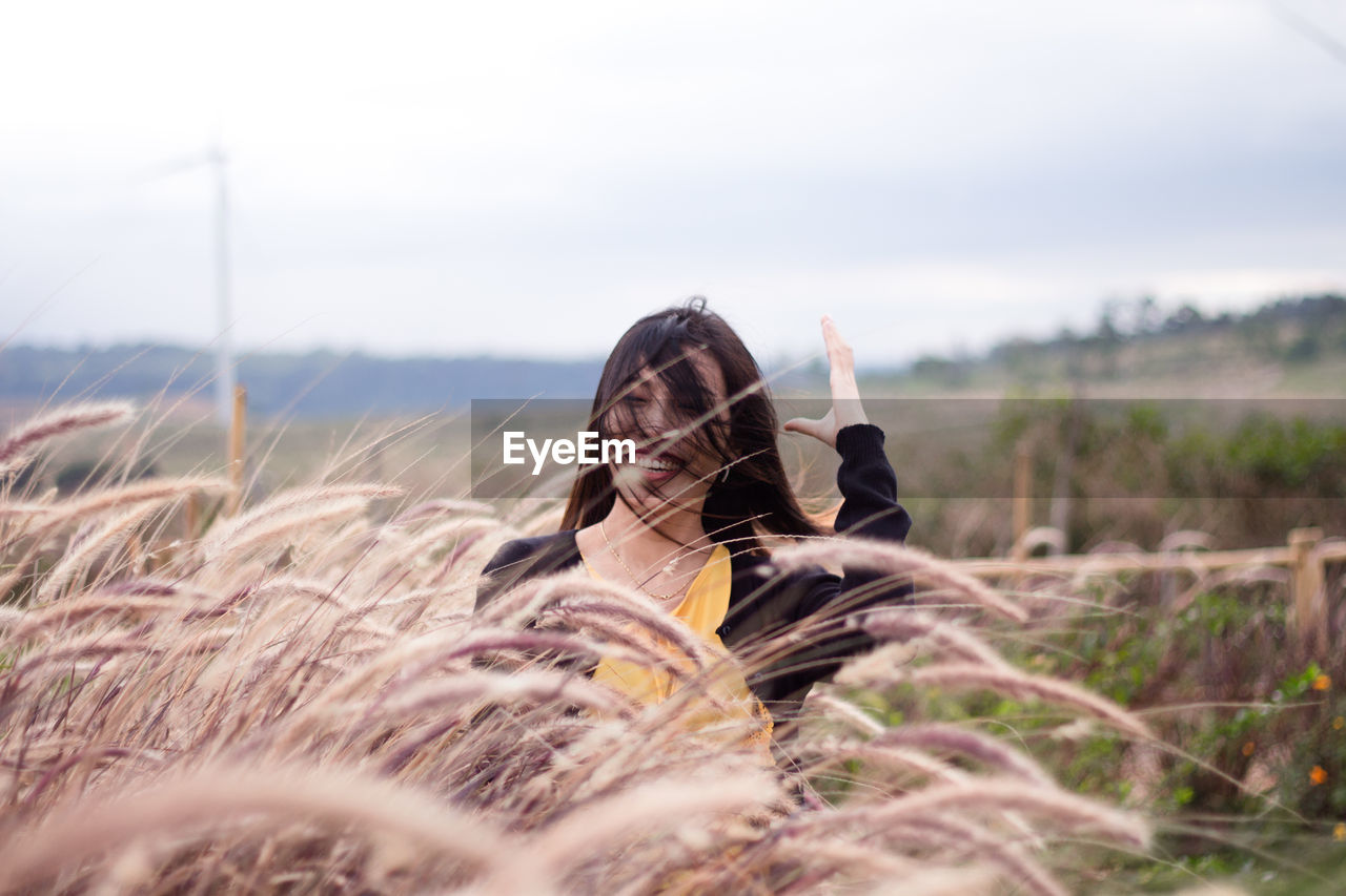 Cheerful woman with tousled hair amidst plants on field against sky