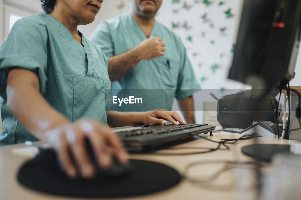 Midsection of nurse using computer by colleague at reception desk in hospital