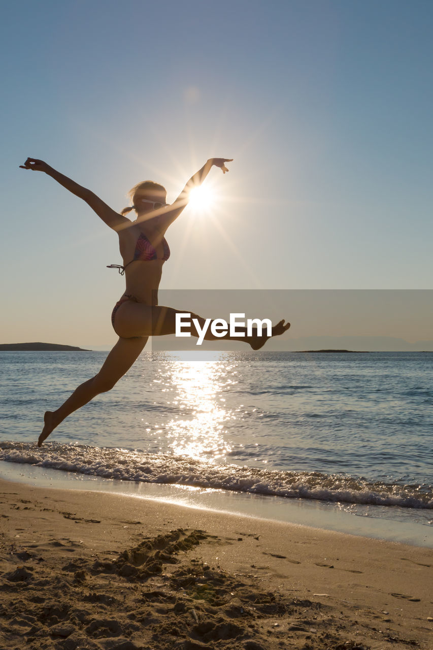 Full length of woman in bikini jumping on beach against sky during sunset