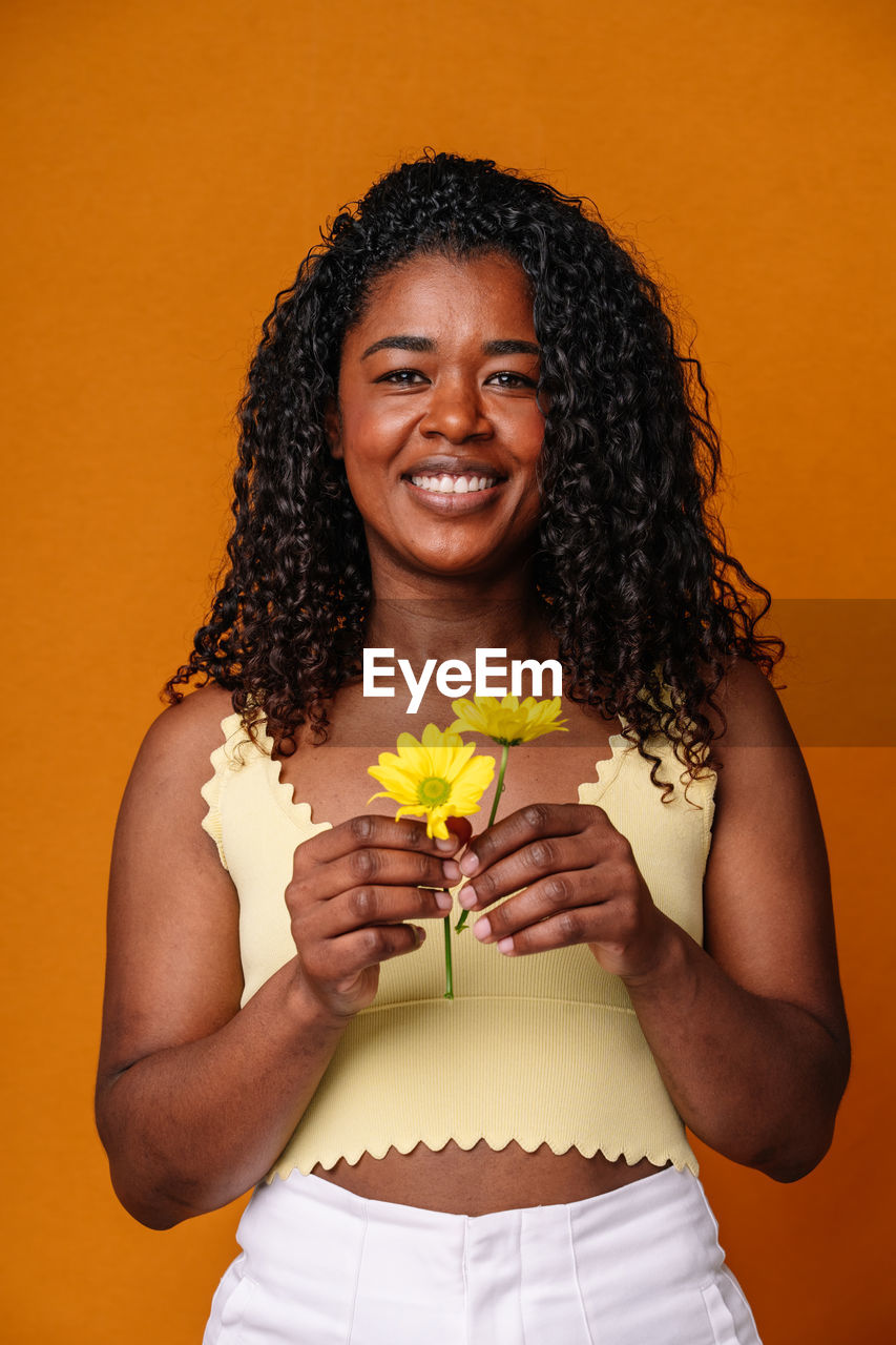 portrait of young woman holding yellow flower against orange background