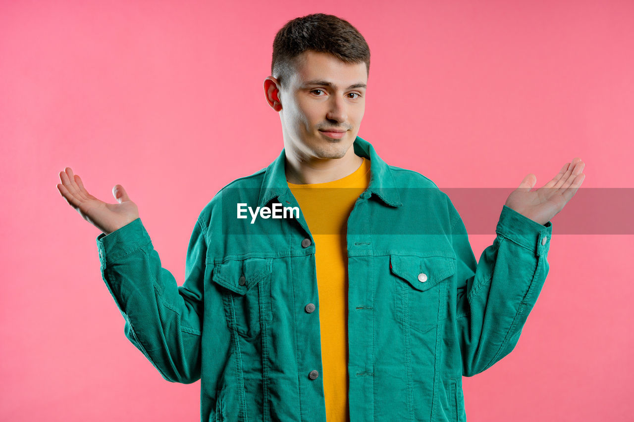 studio shot, one person, colored background, young adult, portrait, waist up, adult, outerwear, looking at camera, standing, indoors, hood, front view, men, pink, clothing, green, jacket, pink background, sleeve, emotion, casual clothing, spring, gesturing