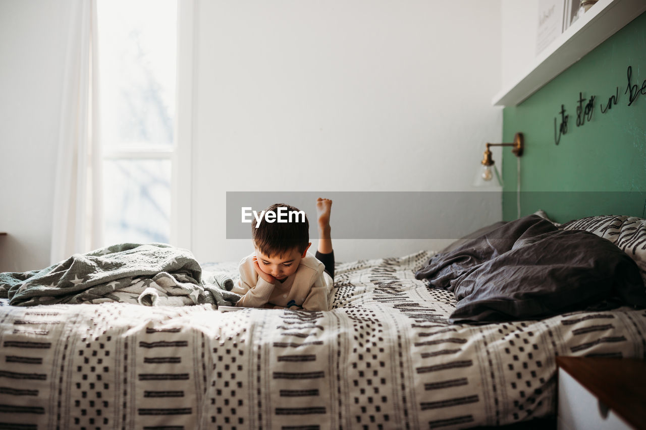 Young boy laying on bed watching on tablet in modern bedroom
