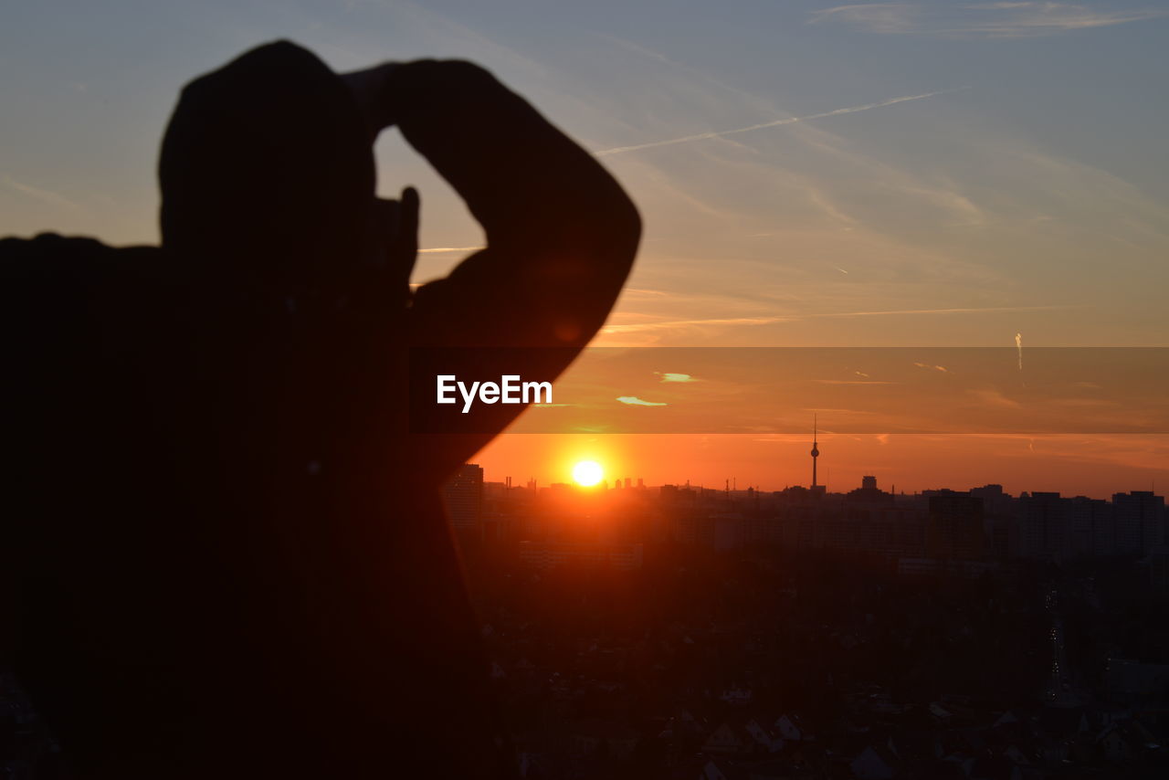 sky, silhouette, sunset, sunlight, one person, nature, backlighting, architecture, technology, evening, horizon, orange color, dawn, building exterior, adult, city, lifestyles, cloud, built structure, photographing, outdoors, sun, cityscape, wireless technology, light, communication, leisure activity, activity, smartphone, men, holding, hand, back lit