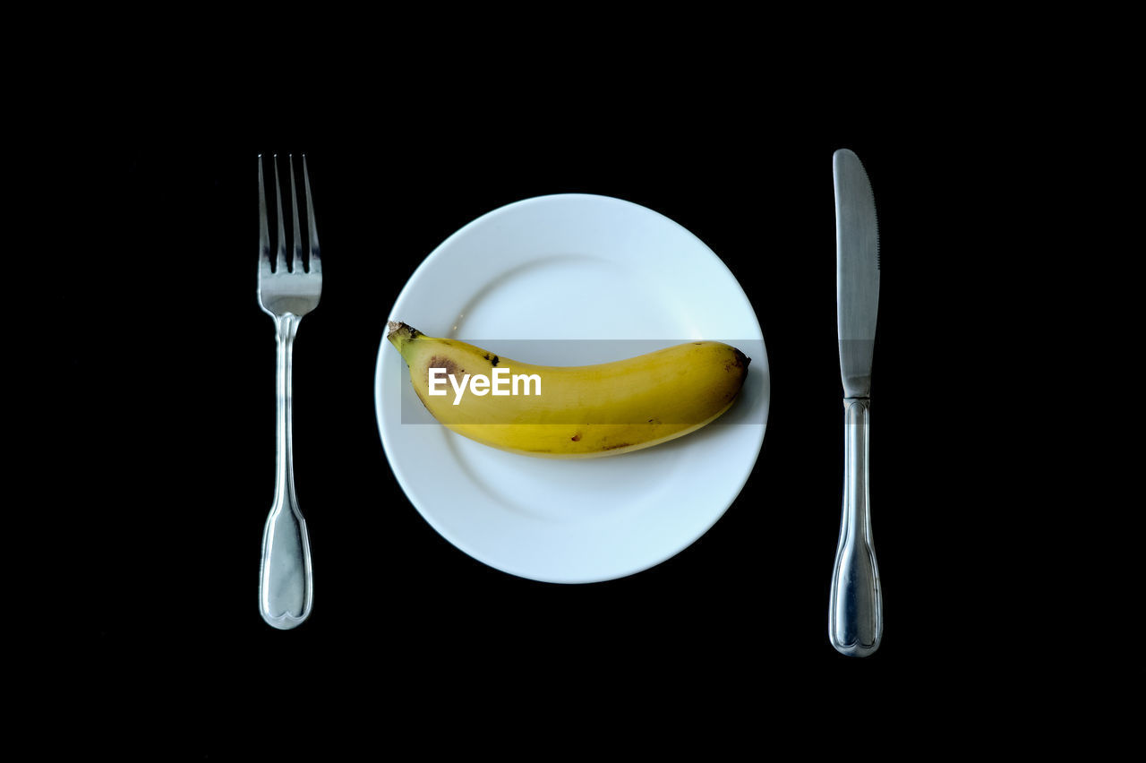 Directly above shot of banana in plate with fork and knife against black background