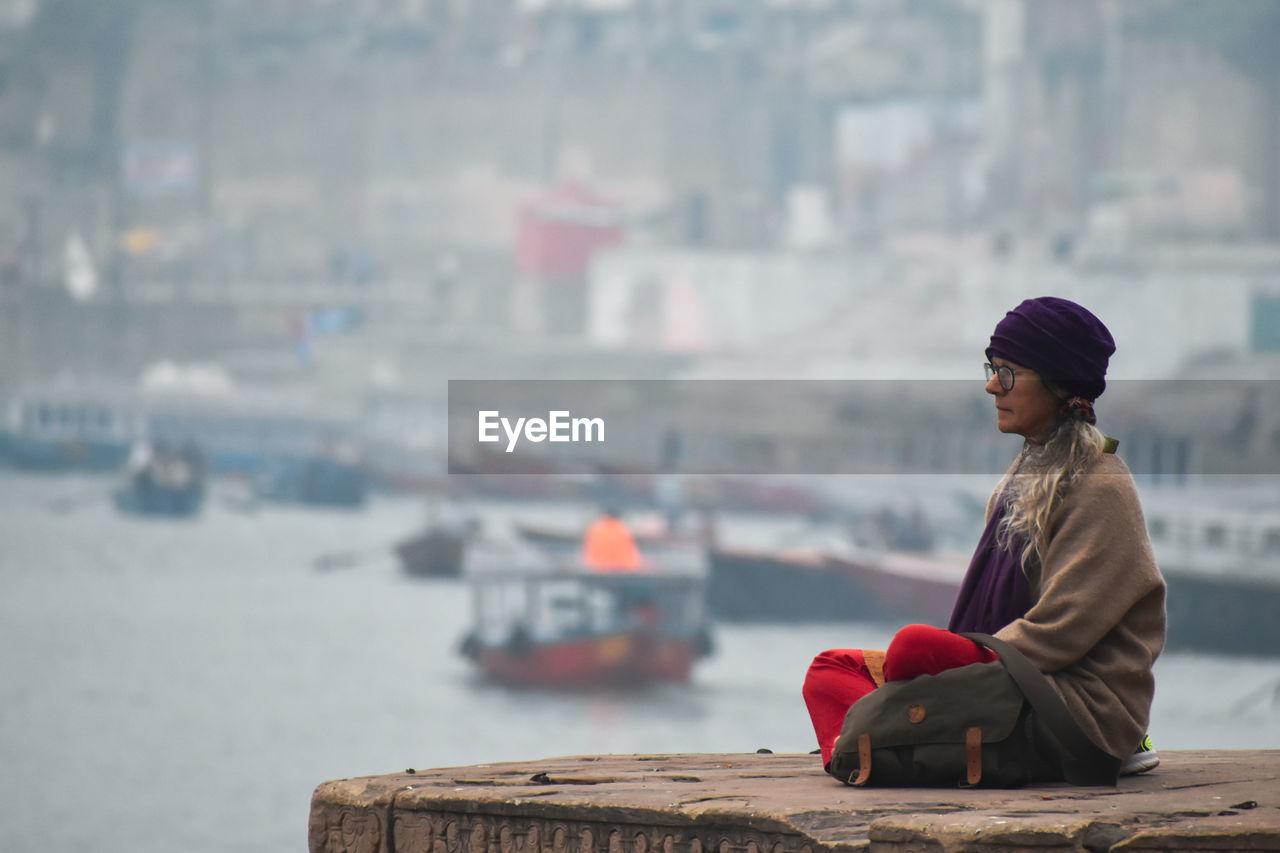 REAR VIEW OF WOMAN SITTING AGAINST BLURRED WATER