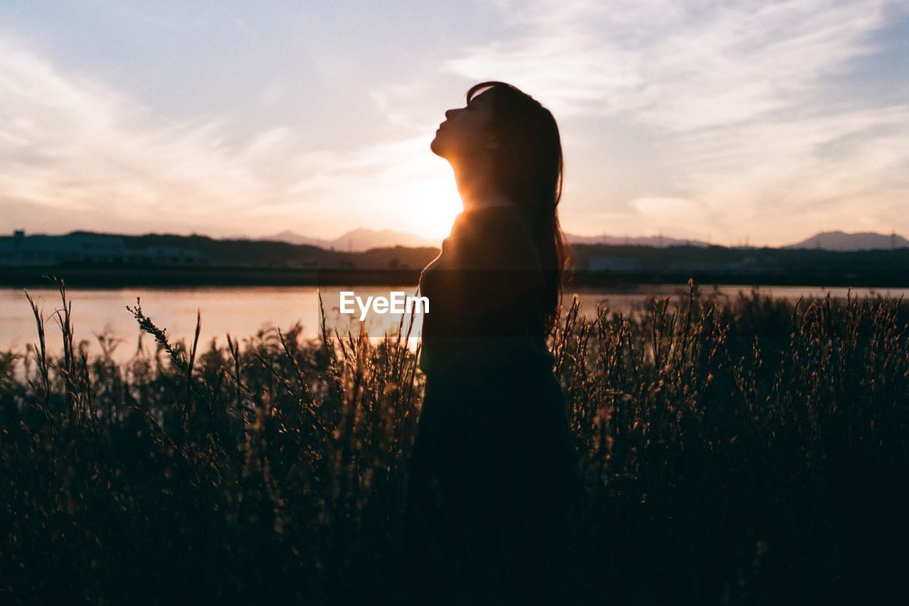 REAR VIEW OF WOMAN STANDING ON FIELD AGAINST SKY DURING SUNSET