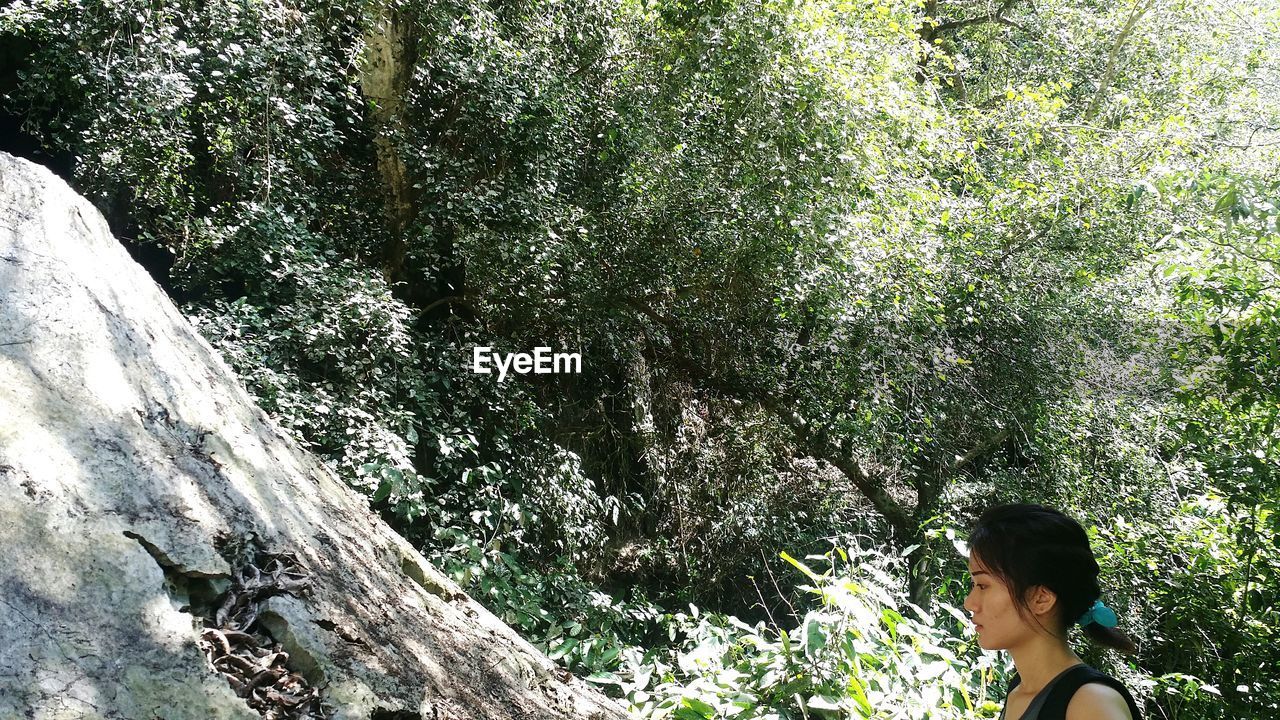 Young woman looking away standing by rock formation against plants