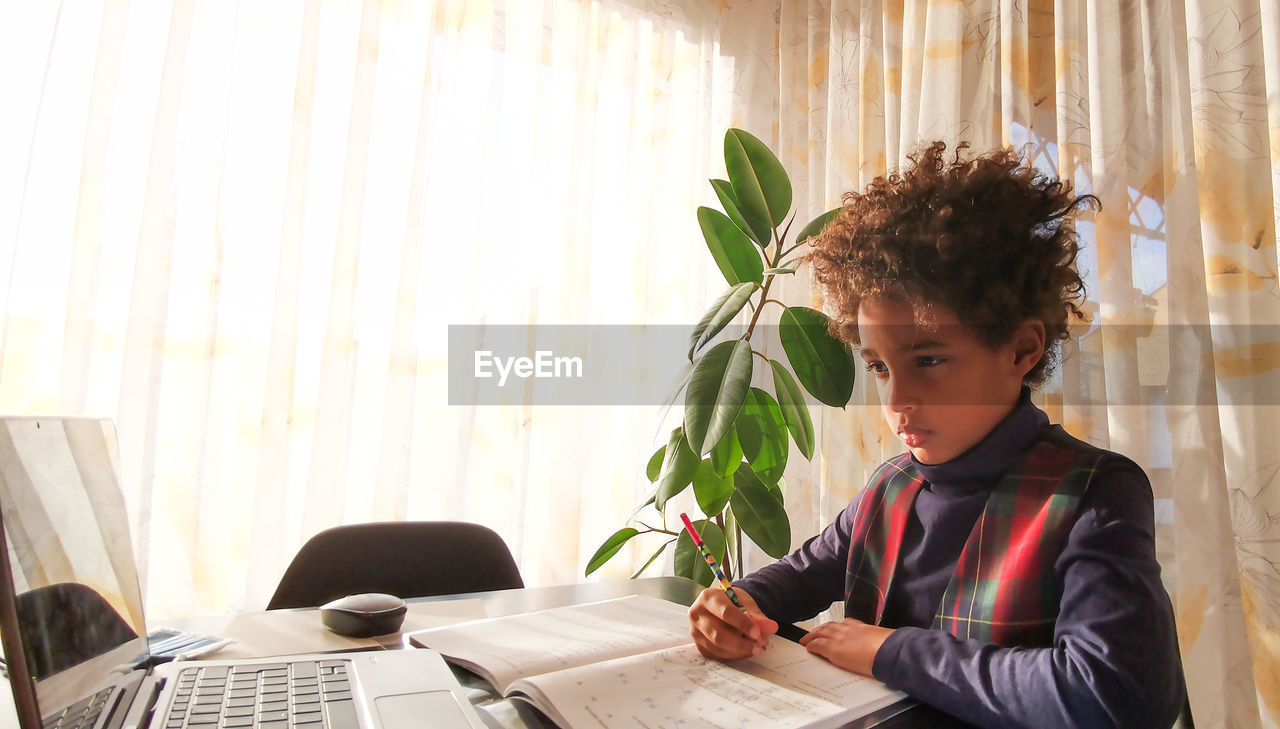 Mixed race kid with curly hair doing remote homework in front of laptop