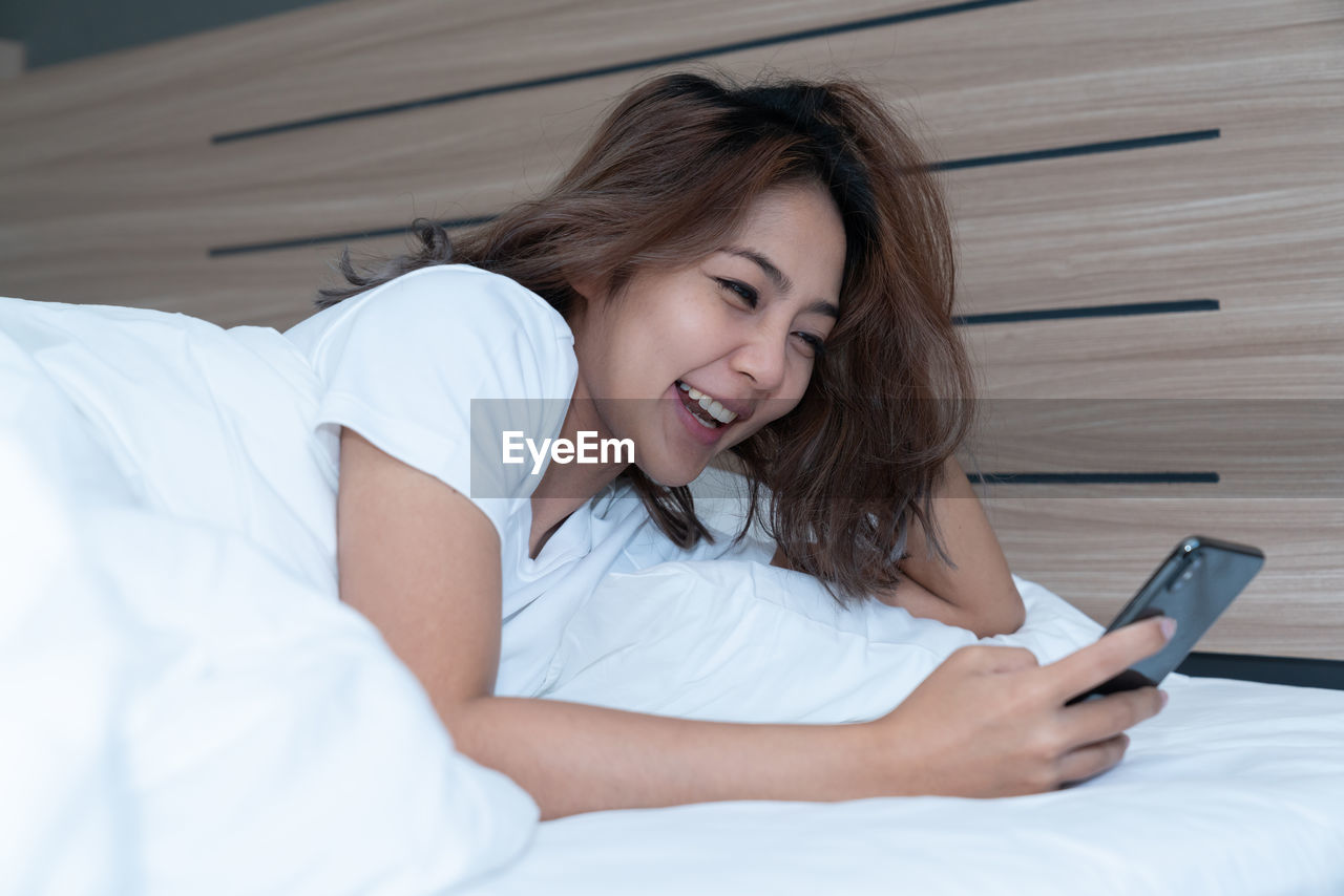 Smiling woman using mobile phone while lying down on bed at home