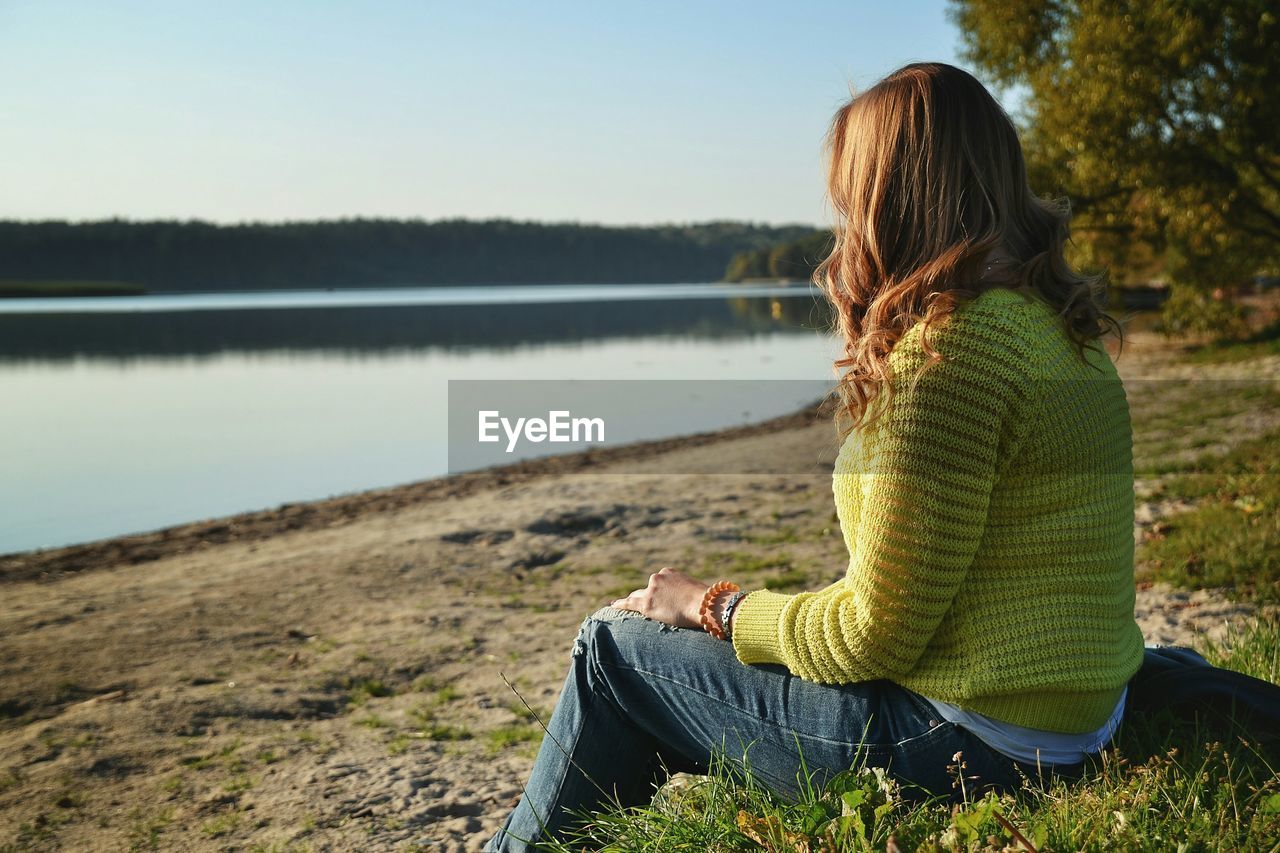 Side view of woman sitting on field by lake during sunny day