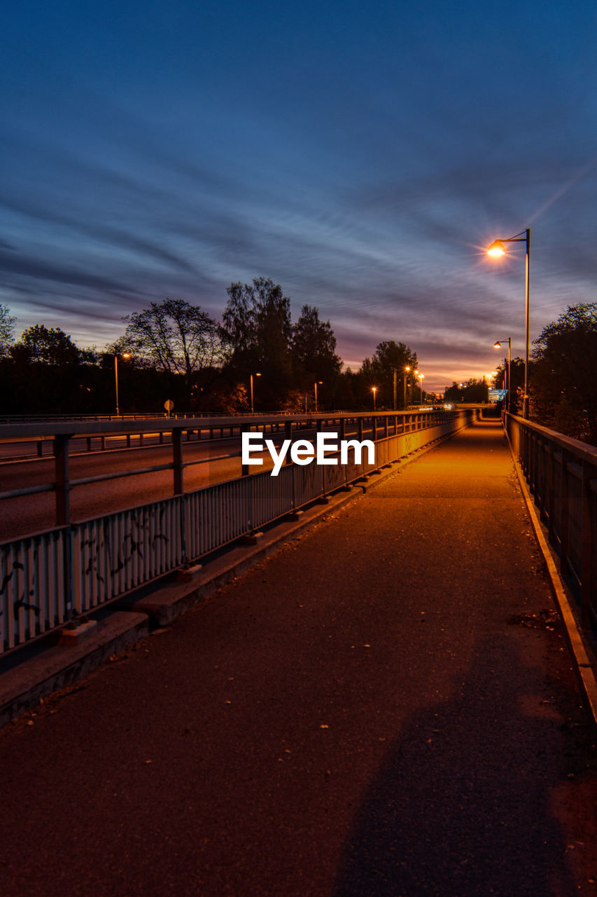 Road by illuminated street against sky at sunset