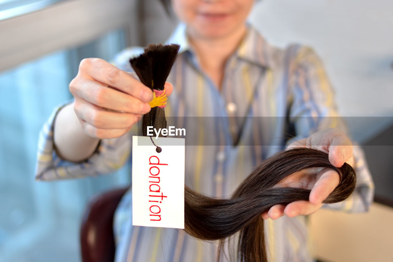 Long brown cut tail with tag in female hands, woman donation of haircut, depth of field