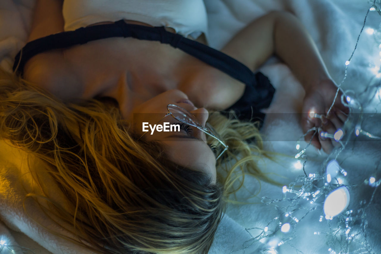 Close-up of woman with illuminated string light lying on bed 
