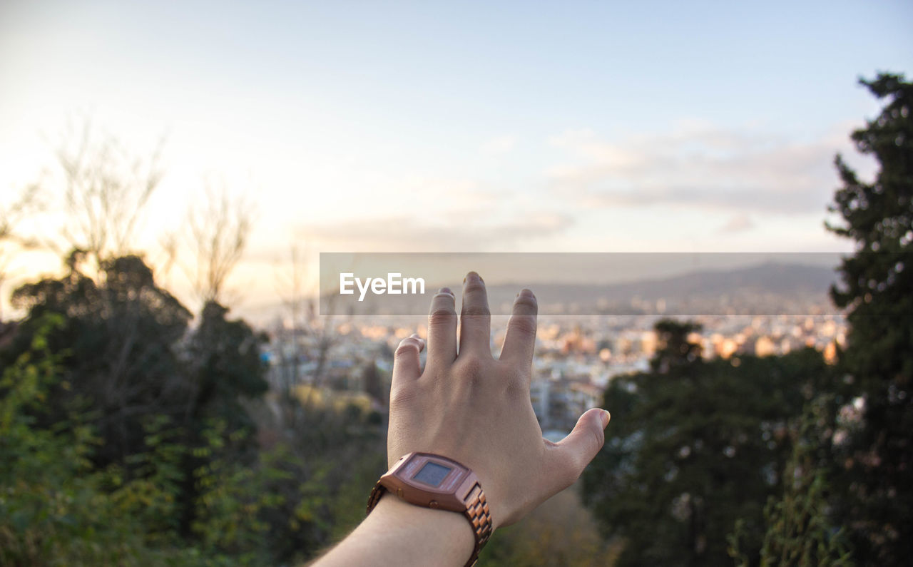 Cropped image of hand against residential district and sky during sunset