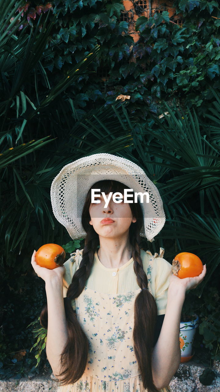 Thoughtful young woman holding fruits while standing against plants