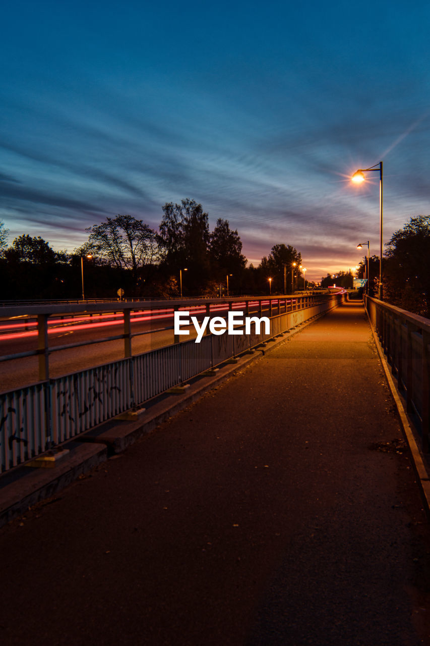 Road by illuminated city against sky at sunset