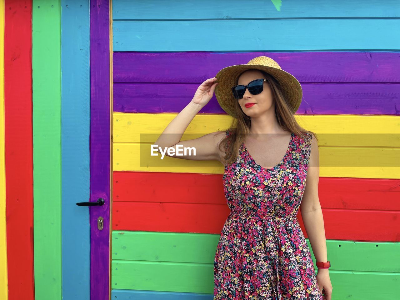 Fashionable young woman wearing straw hat and sunglasses standing against bright and colorful wall