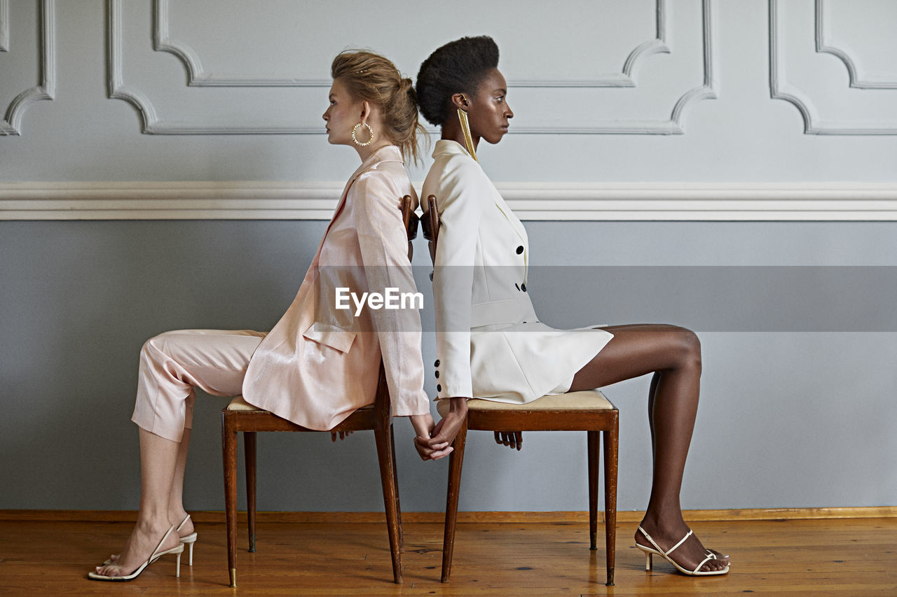 Side view of women holding hands while sitting on chair against wall