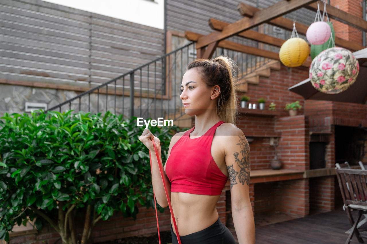 Cheerful female athlete in sportswear doing bicep curl exercise with elastic band during workout in courtyard and looking away