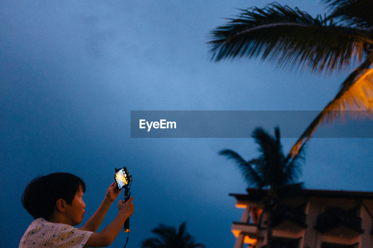 Side view of boy photographing from mobile phone on monopod against sky