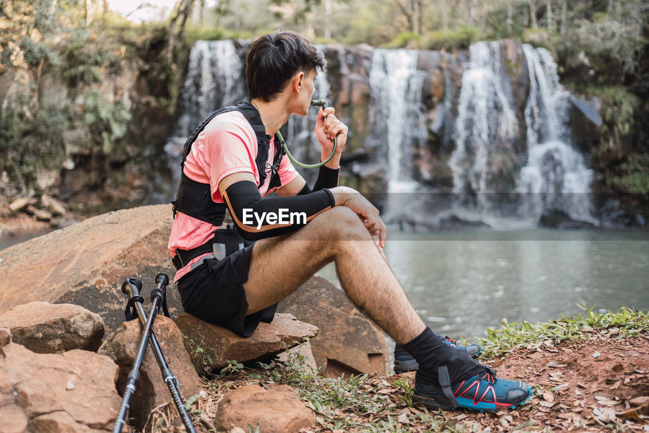 Side view of male hiker with hydration pack drinking from hose while having break during trekking in forest and admiring view of lake near waterfall