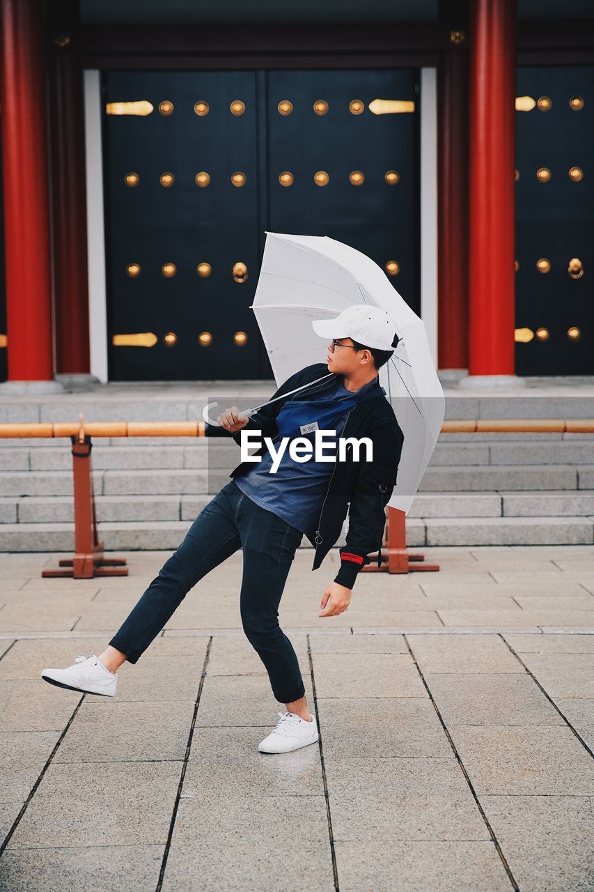 Young man holding umbrella while standing on footpath in city