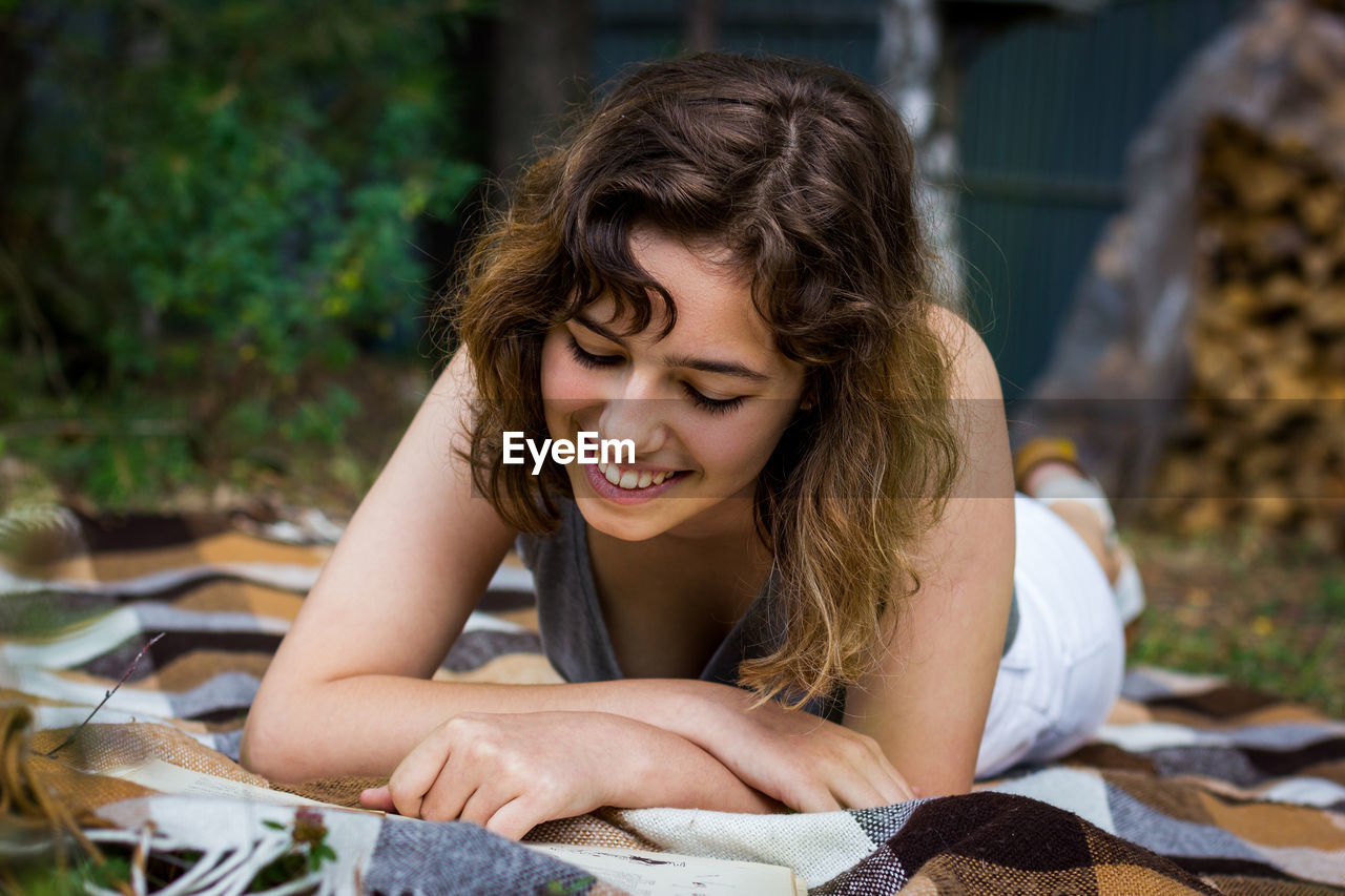 Beautiful teenager girl reading a book lying on blanket on green grass at the woods.