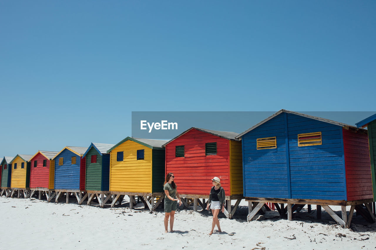 Friends at beach against colorful huts and clear blue sky during sunny day
