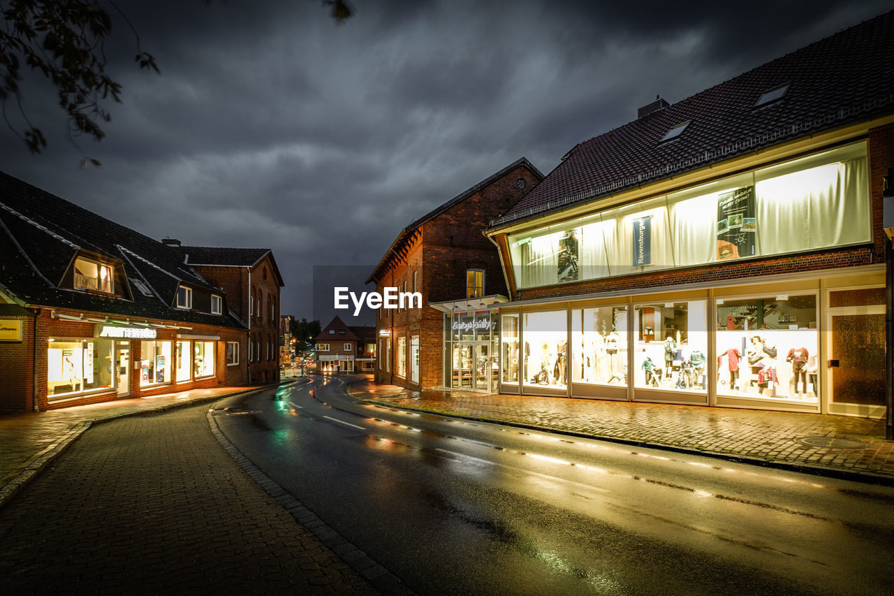 Wet street amidst illuminated stores against cloudy sky at night