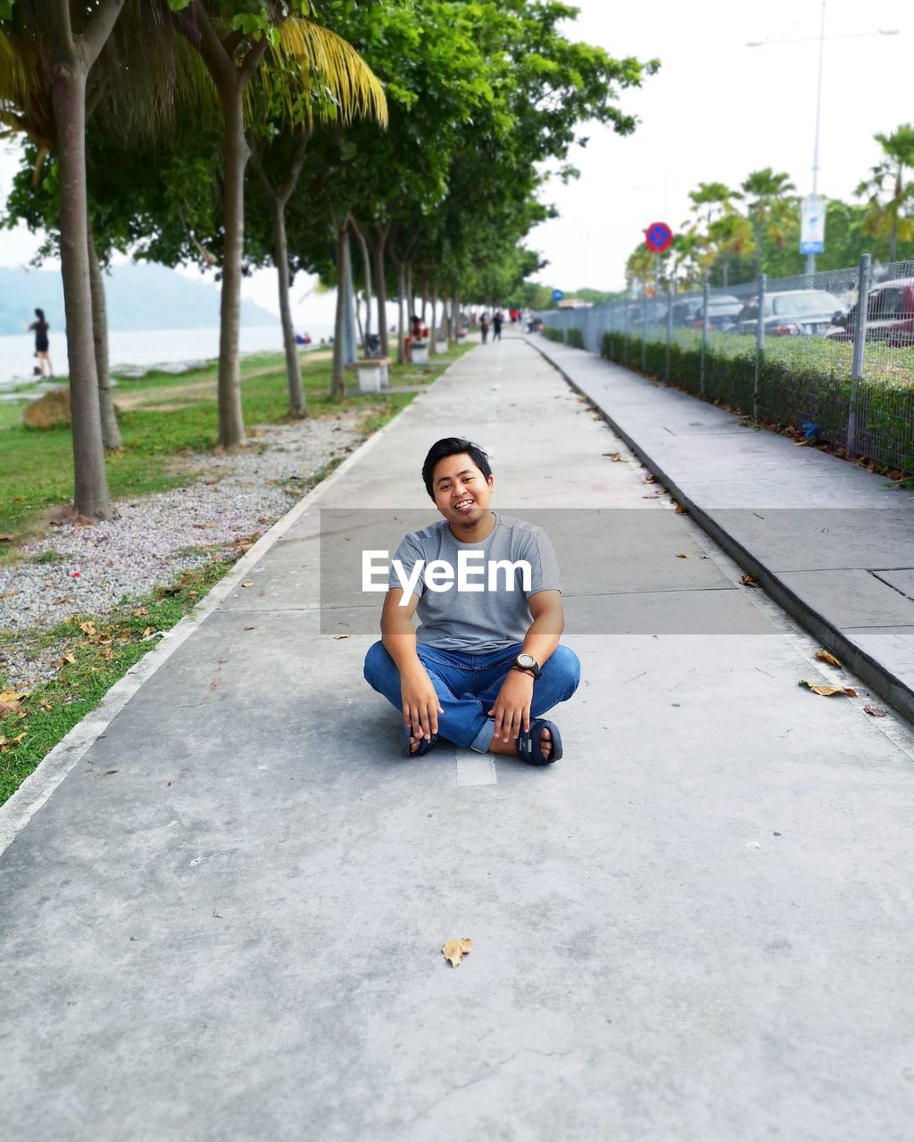 Portrait of young man sitting on sidewalk against trees