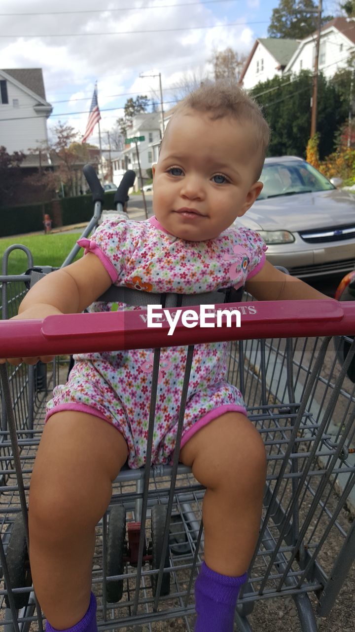 Portrait of baby girl sitting in shopping cart on ground against sky